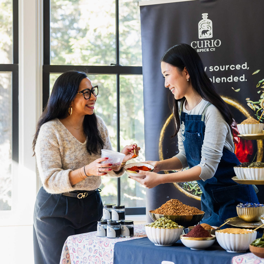 Two women at a Curio Spice Co. event, one holding a bowl of food, with a branded backdrop.