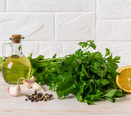 Bottle of olive oil, fresh parsley, lemon, garlic, and peppercorns on a white surface with a white tiled wall background.

