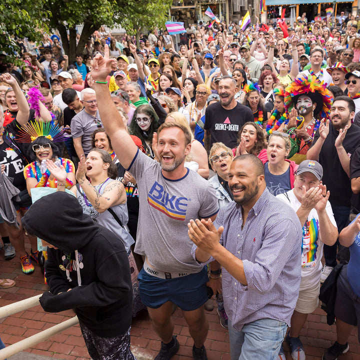 Large crowd of people at a public event, some wearing rainbow colors, with a man in a &