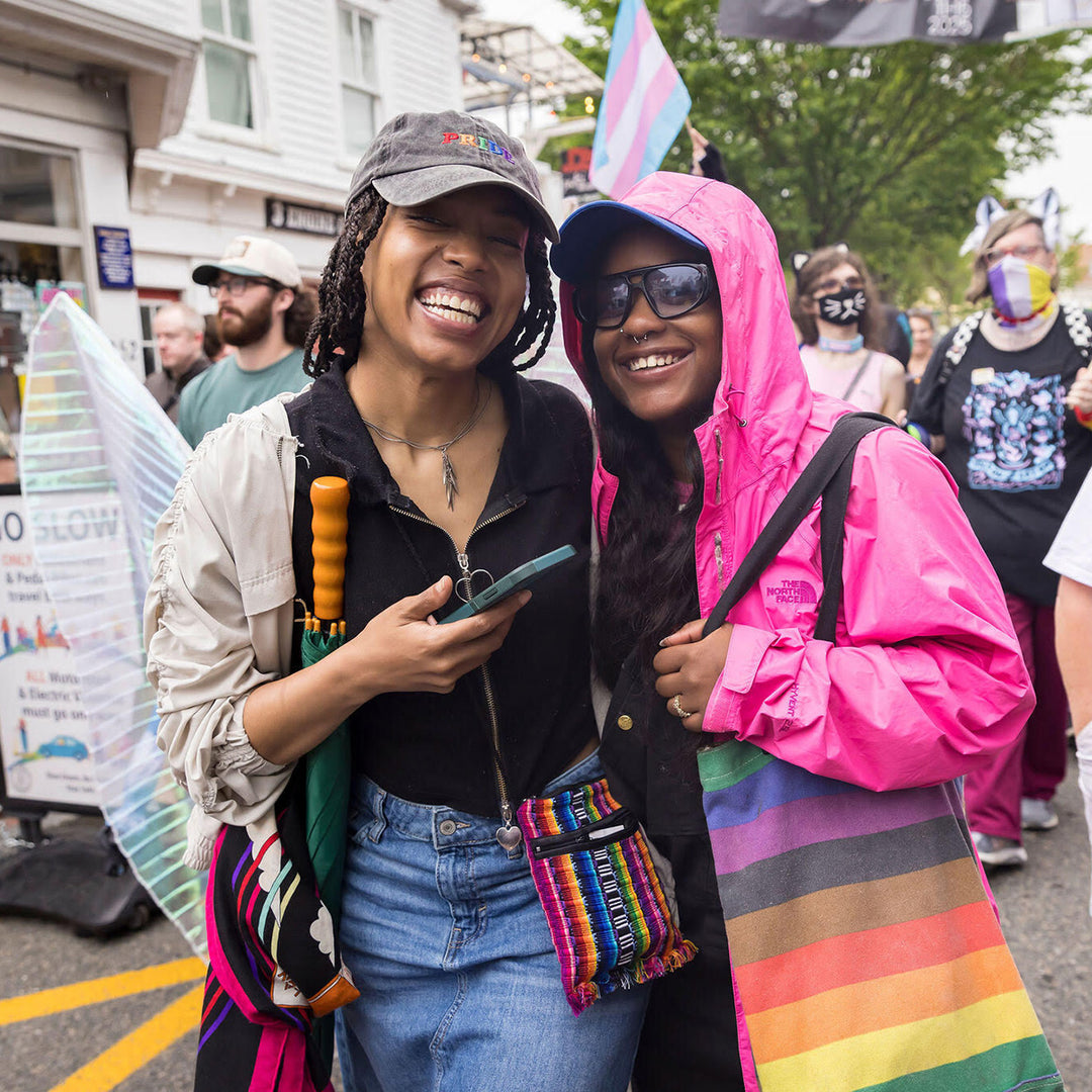 Two people smiling at a protest or rally with rainbow flags and accessories.