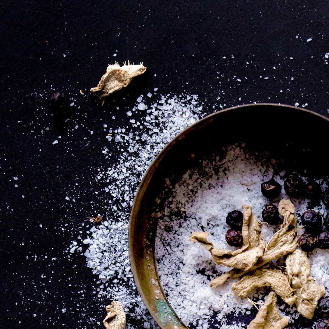 Spices including dried roots and berries on a bed of salt in a bowl against a dark background