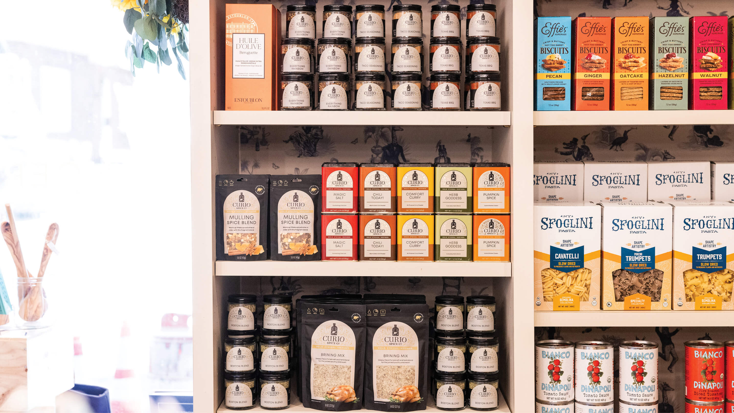 A white wooden shelf stocked with various packaged food products, including jars, boxes, and cans of spaghetti and pasta from brands like Spogolini and Cafe Italiano.